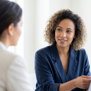 A smiling professional woman in a blazer gestures while discussing financial matters with a client in a bright office.